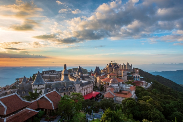 Vue panoramique d'un village de château de style européen sur une colline au coucher du soleil, entouré d'une verdure luxuriante et de montagnes lointaines, parfait pour les amateurs de voyages et d'architecture.