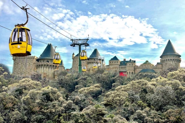 Des téléphériques baptisés « Ba Na Hills » survolent un château de conte de fées doté de multiples tourelles, situé au milieu d'une végétation dense sous un ciel partiellement nuageux, idéal pour l'aventure et le tourisme.