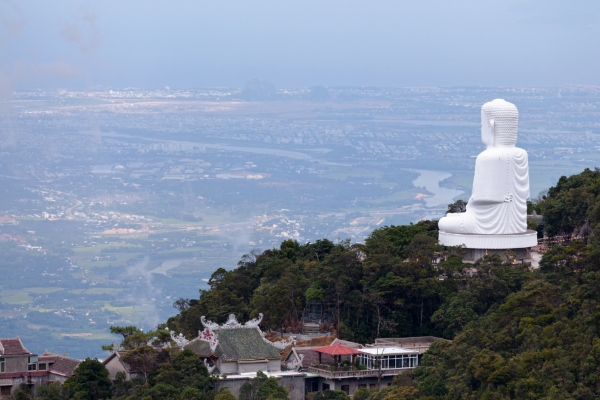 Grande statue de Bouddha blanc sur une colline surplombant une ville et une rivière, entourée d'une verdure luxuriante et de bâtiments traditionnels, idéale pour les voyages et le tourisme spirituel.