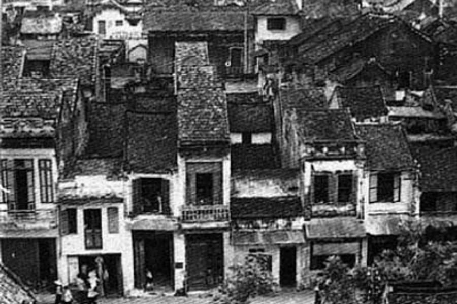 A black and white photograph of Hanoi's Old Quarter. A row of old buildings huddled together with tiled roofs and boarded-up windows.