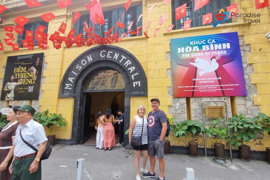 Un couple profitant d'une visite culturelle à la Maison Centrale (la prison de Hoa Lo) à Hanoi, debout devant le bâtiment jaune historique orné de drapeaux vietnamiens, explorant la riche histoire et les attractions de la ville.