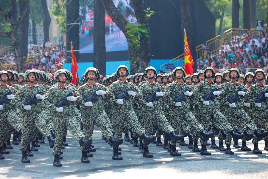 Défilé militaire avec des soldats en uniformes de camouflage marchant à l'unisson, tenant des fusils, avec des drapeaux vietnamiens et une grande foule en arrière-plan, symbolisant la fierté nationale et l'unité.